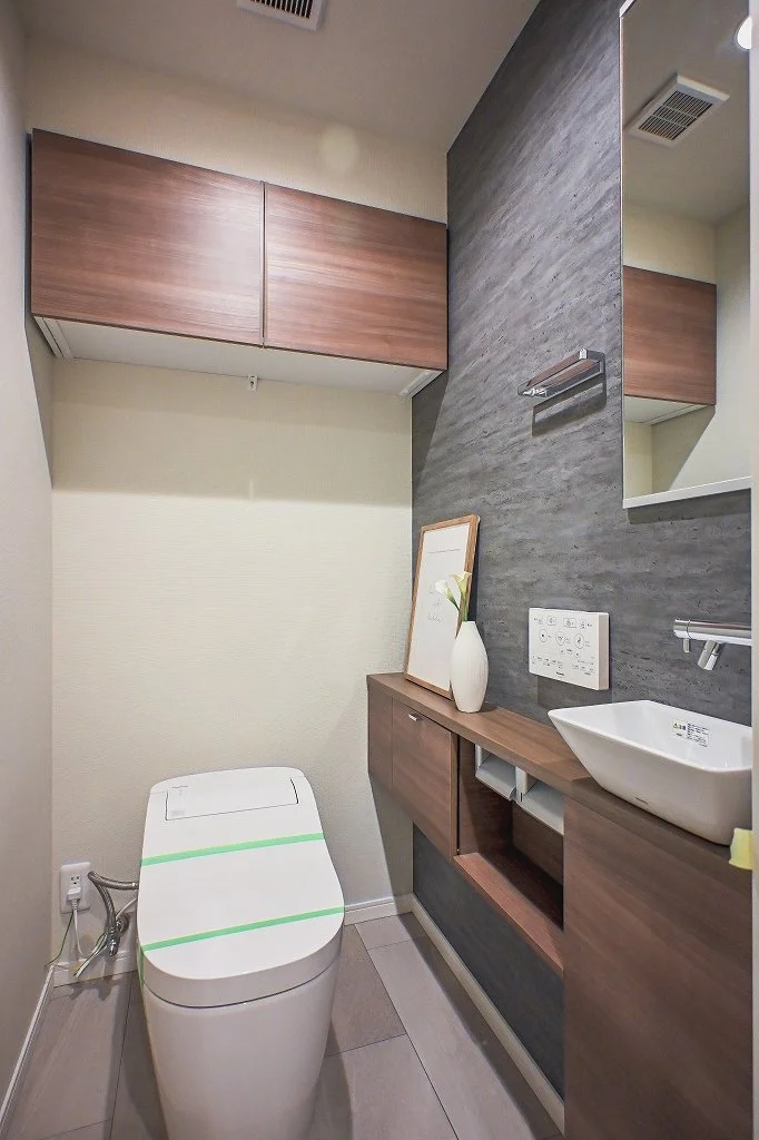 モダンなトイレルーム、木製の収納棚と洗面台、壁に鏡とアクセントクロス。