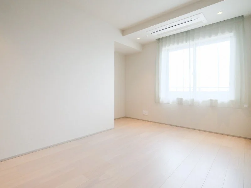 明るい日当たりの良い空き部屋、白い壁と木製の床、窓に白いカーテンがかかっている
