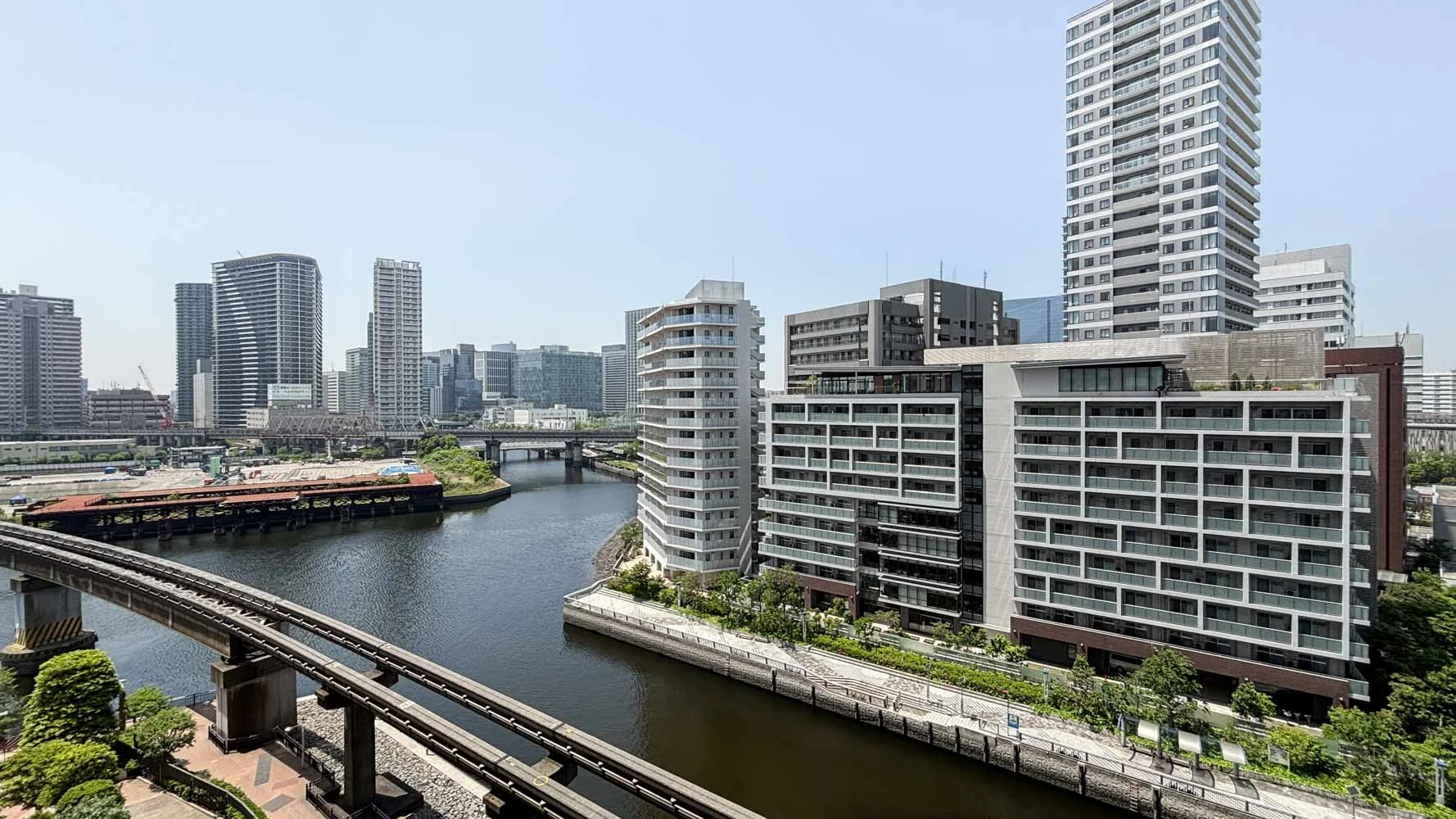都市の高層ビルと川が流れる景色。橋と駅の線路も見える都市風景。
