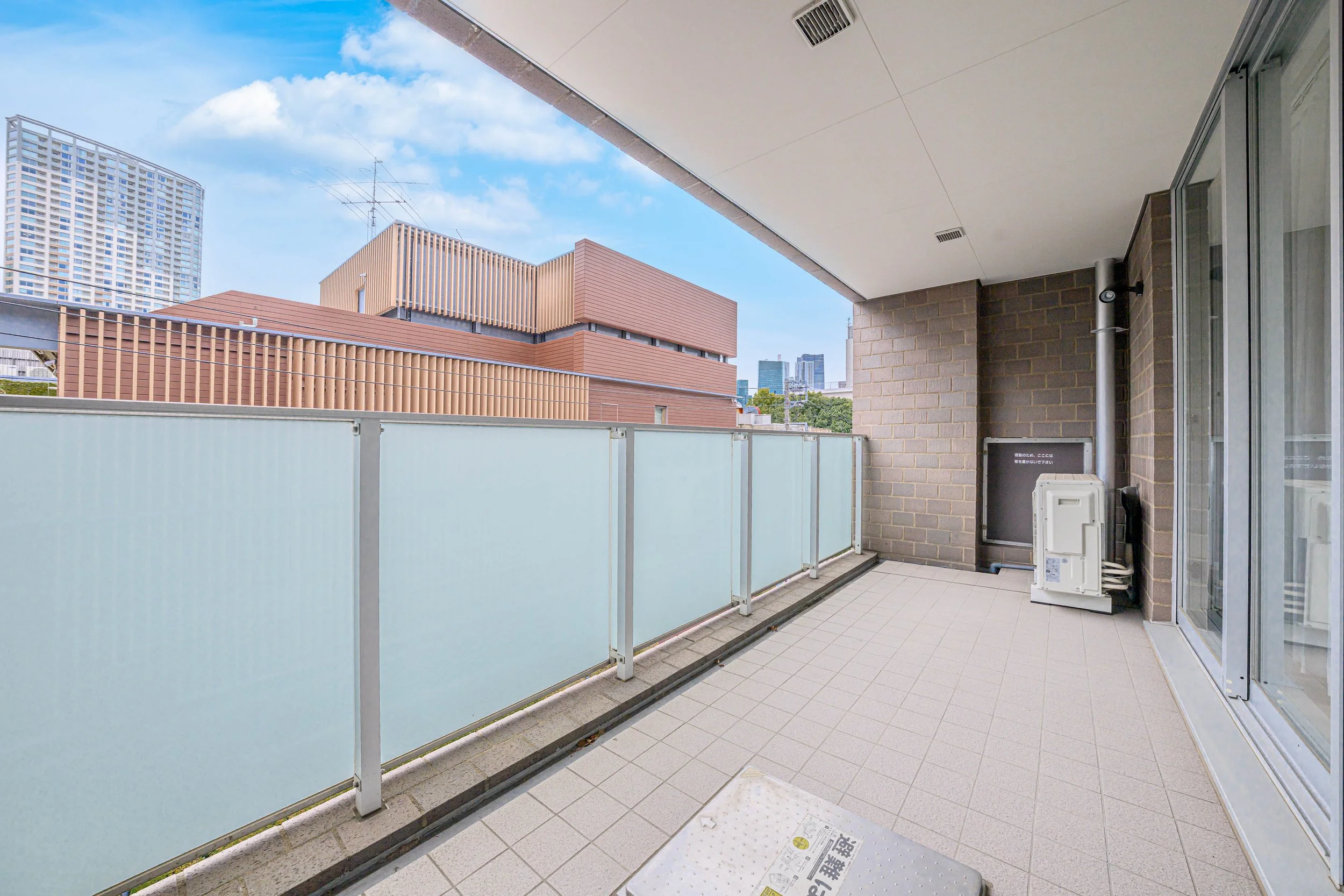 都市の高層ビルの景色が見えるバルコニー。バルコニーにはガラス製の手すりと、空気調節装置らしき白い箱が設置されている。
