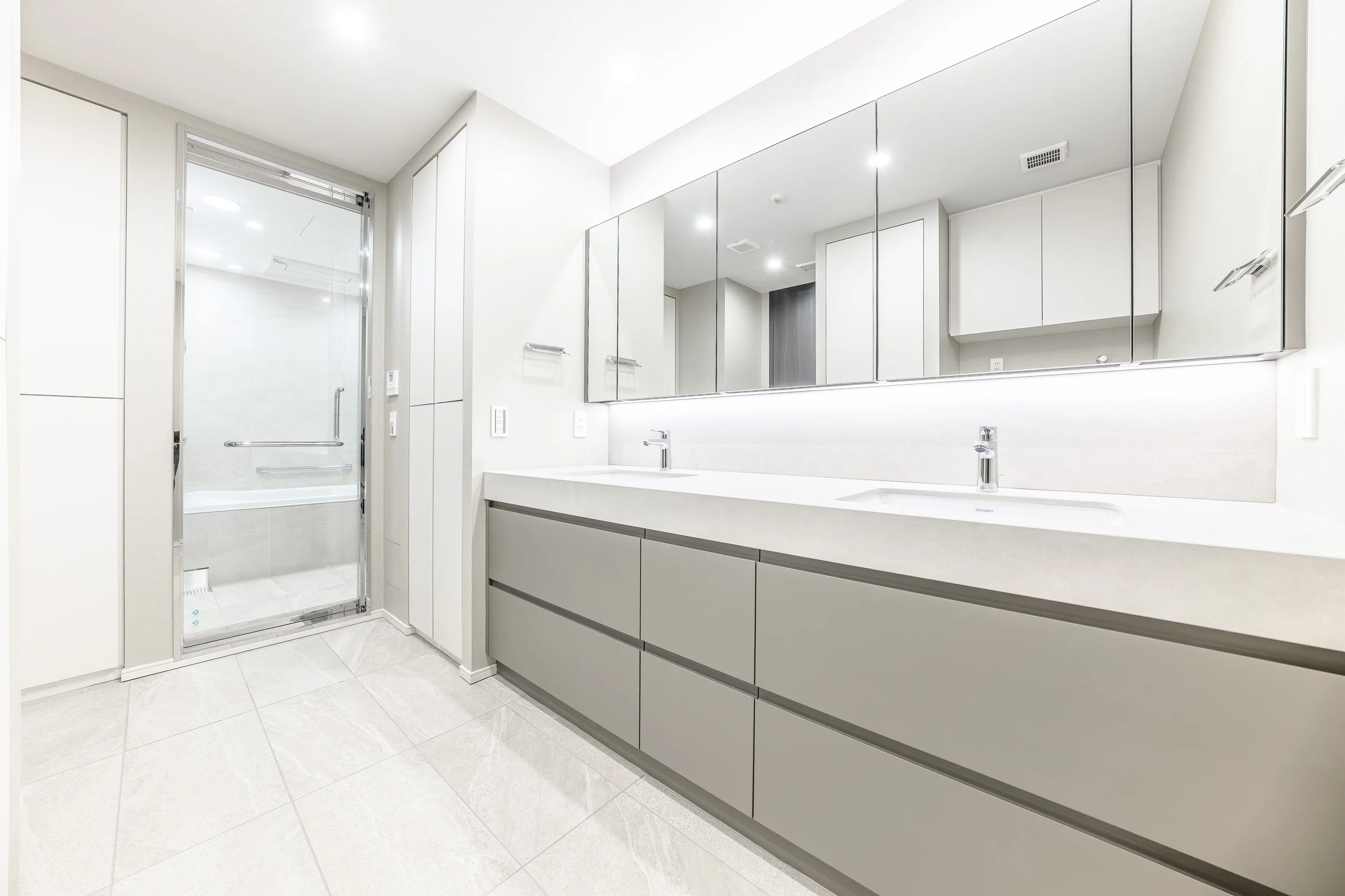 白を基調としたモダンな洗面所と浴室の内部。鏡張りの棚、二つの洗面台とシンプルな収納キャビネット、シャワードア付きのバスタブが見える。