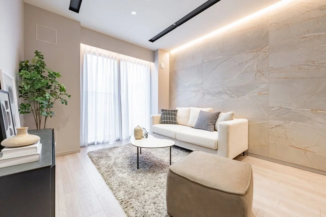 モダンなリビングルームで、白いソファと丸い白いテーブル、グレーのラグとクッションが置かれている。窓には白いカーテンが掛かっており、緑の観葉植物と装飾品も見える。