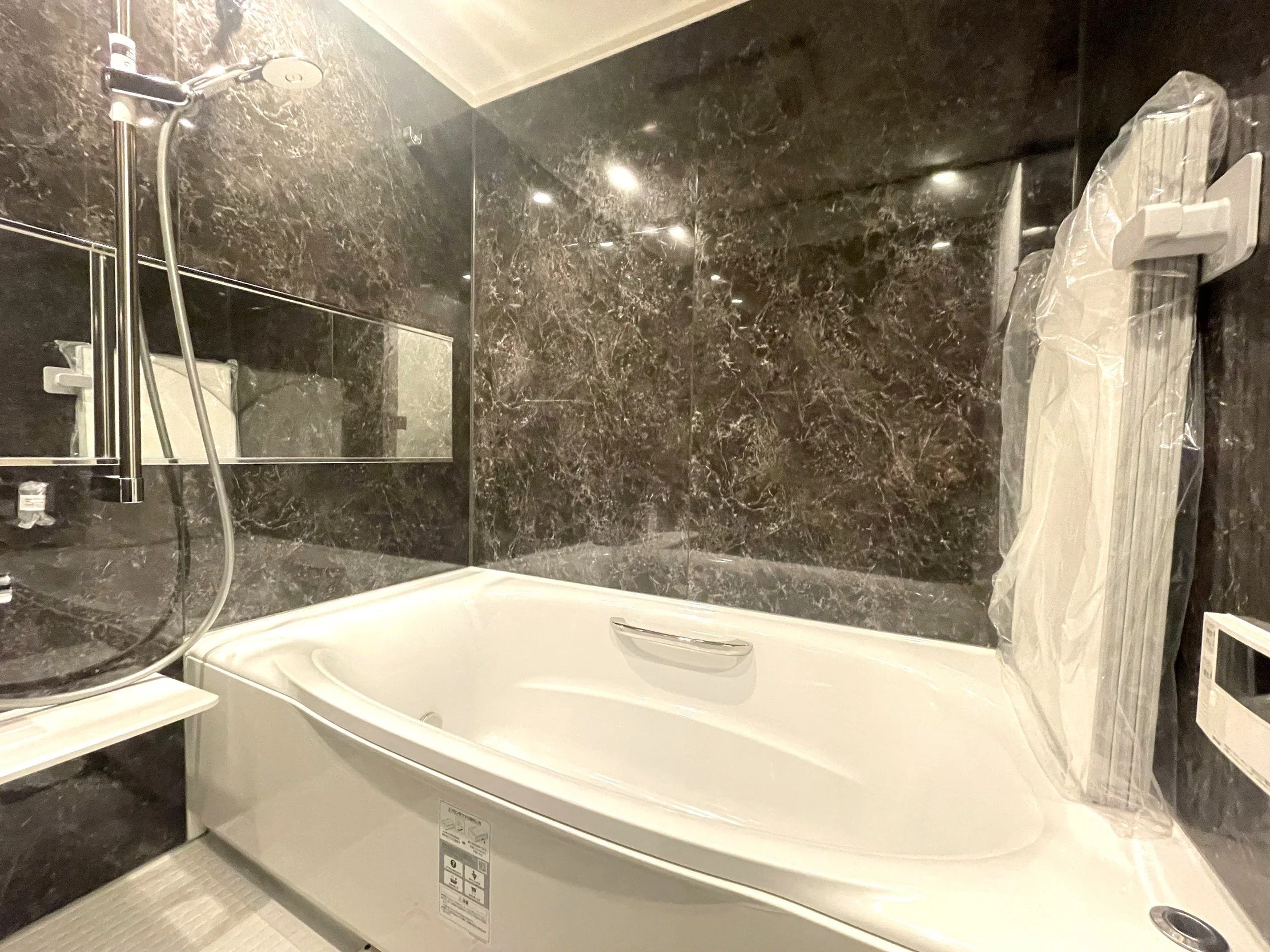 黒と白の大理石柄の壁とバスルームの浴槽。シャワーと鏡も見える。