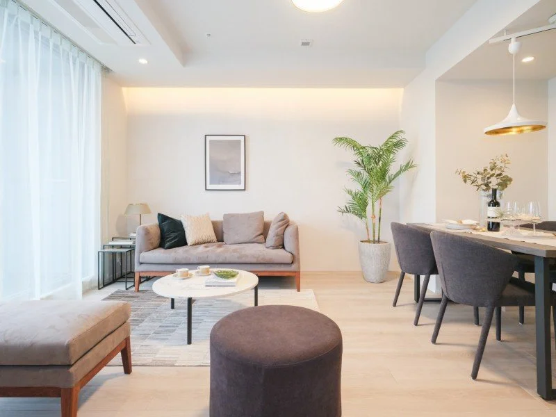 明るいリビングルームで、灰色のソファと丸いコーヒーテーブル、グレーのクッション、観葉植物と飾りが置かれたダイニングエリアが見える。