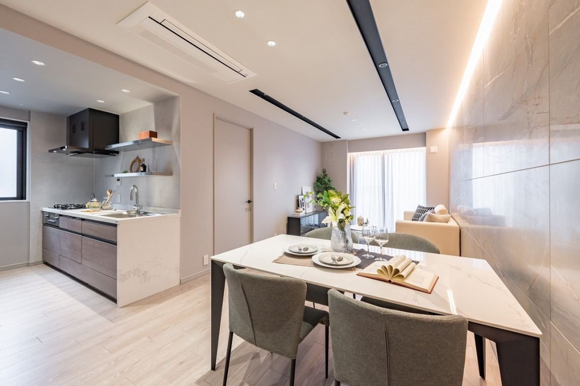 モダンなリビングルームとキッチンのインテリア。ダイニングテーブルに花と食器、グラス、開いた本が置かれている。背後にはソファとテレビ、窓から自然光が差し込む。