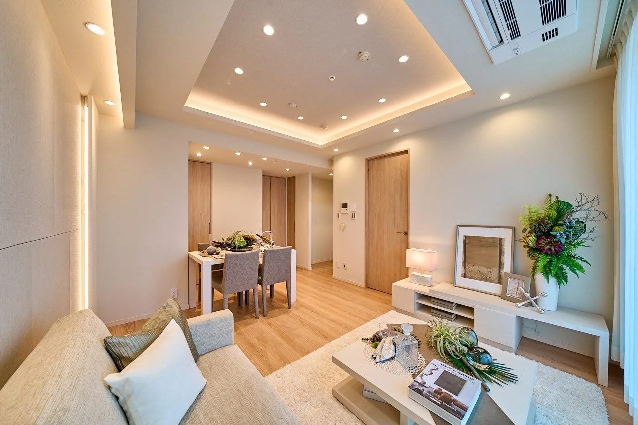 モダンなリビングルームのインテリア写真。ソファ、テーブル、植物、照明、テレビ台などが配置されており、暖色系の照明が落ち着いた雰囲気を作り出している。