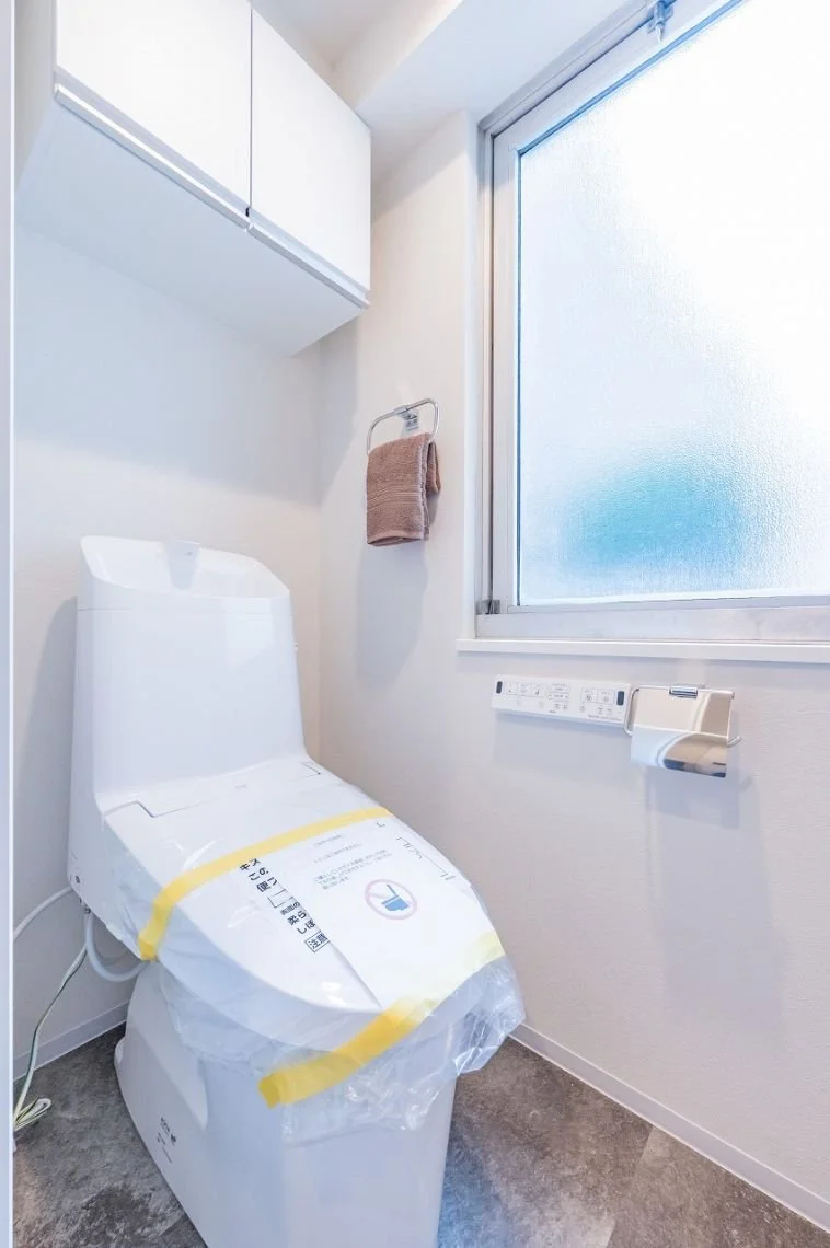 白いトイレとタオル掛け、上部に白い収納棚、横に窓とトイレットペーパーホルダーがあるバスルームの一角