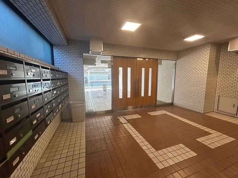 マンションのエントランス、壁に沿って郵便受け箱が並び、中央に木製のドアとガラス扉。その前には掃除用のゴミ箱が置かれている。室内の床にはタイルの模様がある。