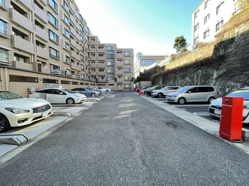 コンクリートの駐車場、複数の車と赤い駐車券リーダー付きの機械が並ぶ都市の一角、背景に高層マンションと岩壁が見える