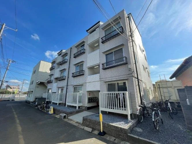 白い多層アパートの建物と自転車が並ぶ駐輪場。青空が背景に広がる風景。