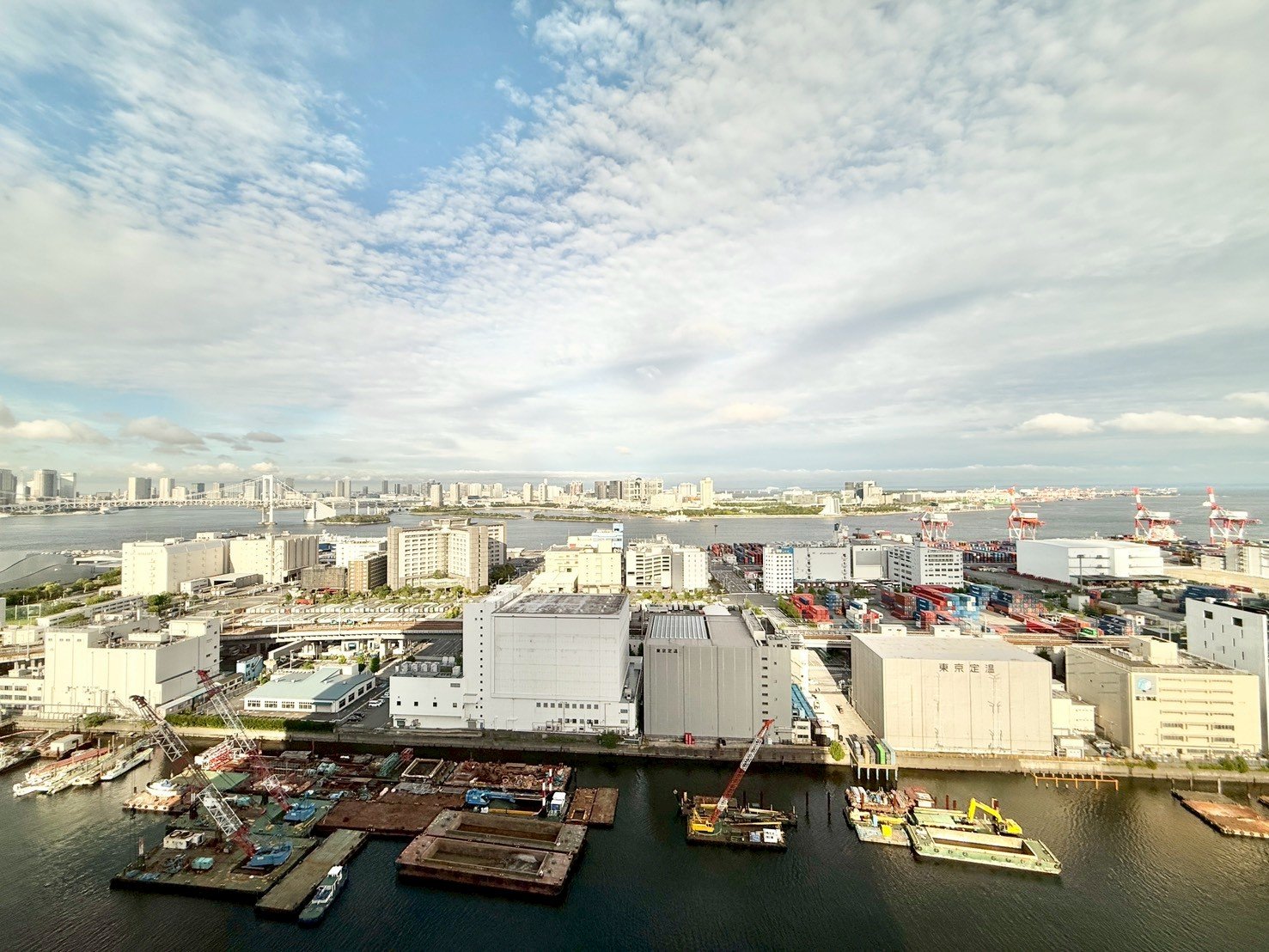 東京の港の景色で、工場や倉庫とクレーンが見える。背後に東京湾と都心の高層ビル群と橋がある。空は曇りと晴れの混合、青空が見える。