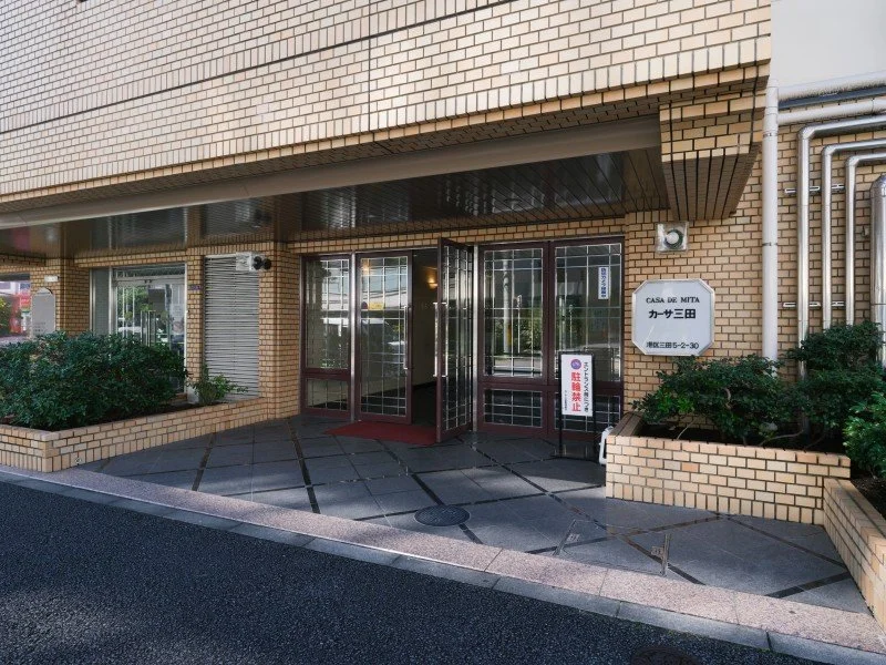 横長のレンガ造りの建物の入口付近。ガラス扉、サインボードと植物が見える。