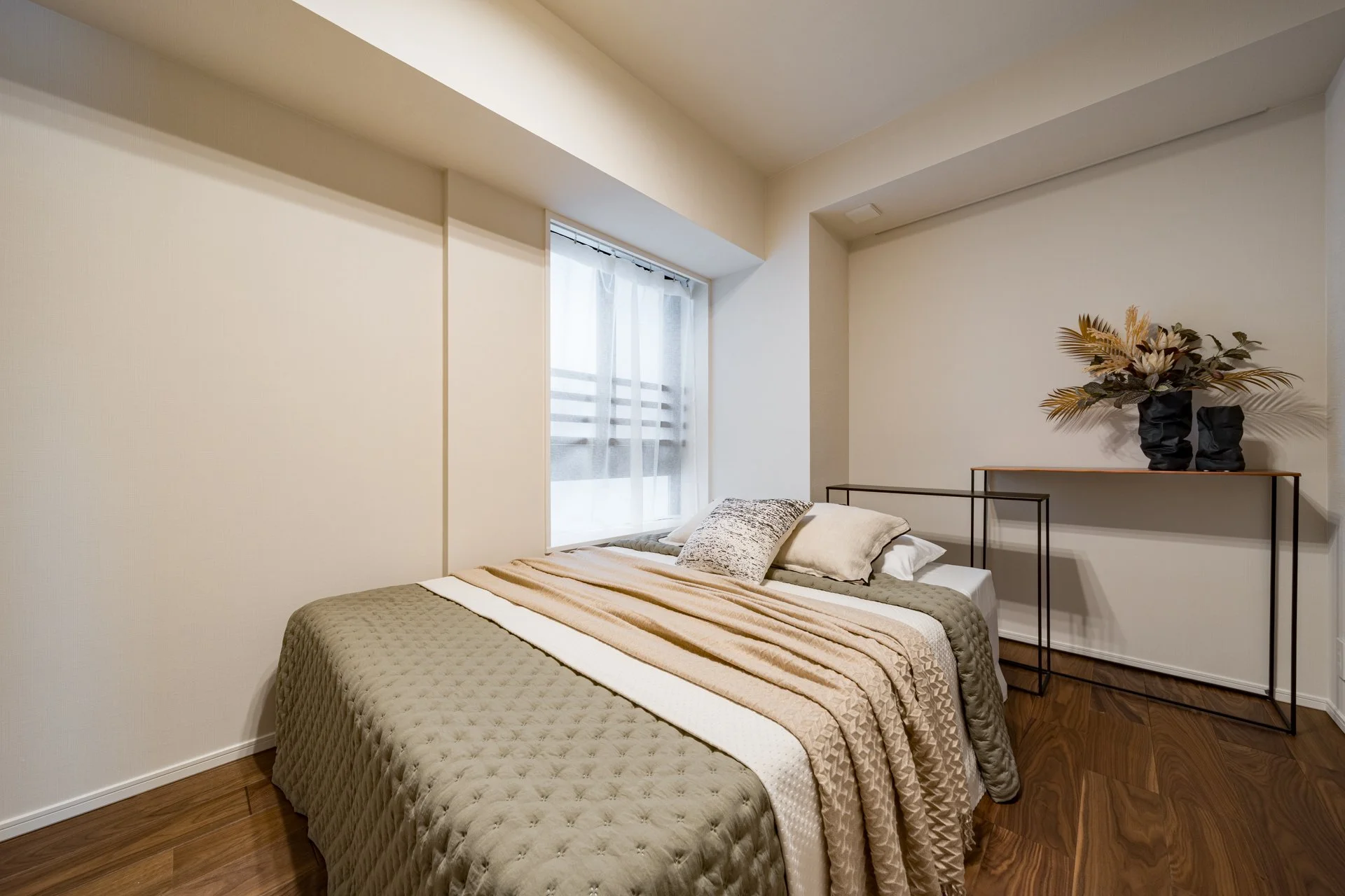 シンプルなベッドルーム。白い壁と木製の床、窓から自然光が差し込み、ベッドにはクッションと掛け布団が置かれている。壁には花の飾りが置かれた黒い陶器の花瓶が置かれた棚がある。