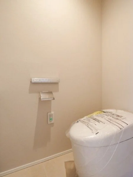 白い壁の部屋にトイレと紙巻き器、棚、リモコンが取り付けられている。