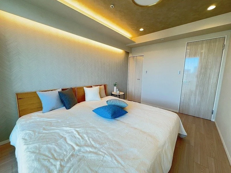 シンプルでモダンな寝室、白いベッドに複数のクッションと枕、木製のサイドテーブルに花瓶と本棚、木のフローリングとシンプルな壁、間接照明のある天井。