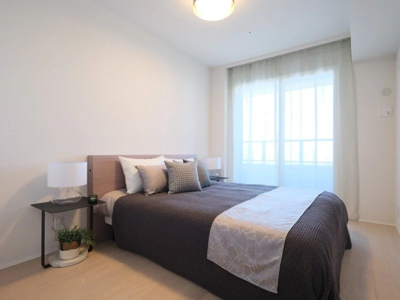 白い壁の部屋にダブルベッドと二つのサイドテーブル、窓には白いカーテンがかかっている