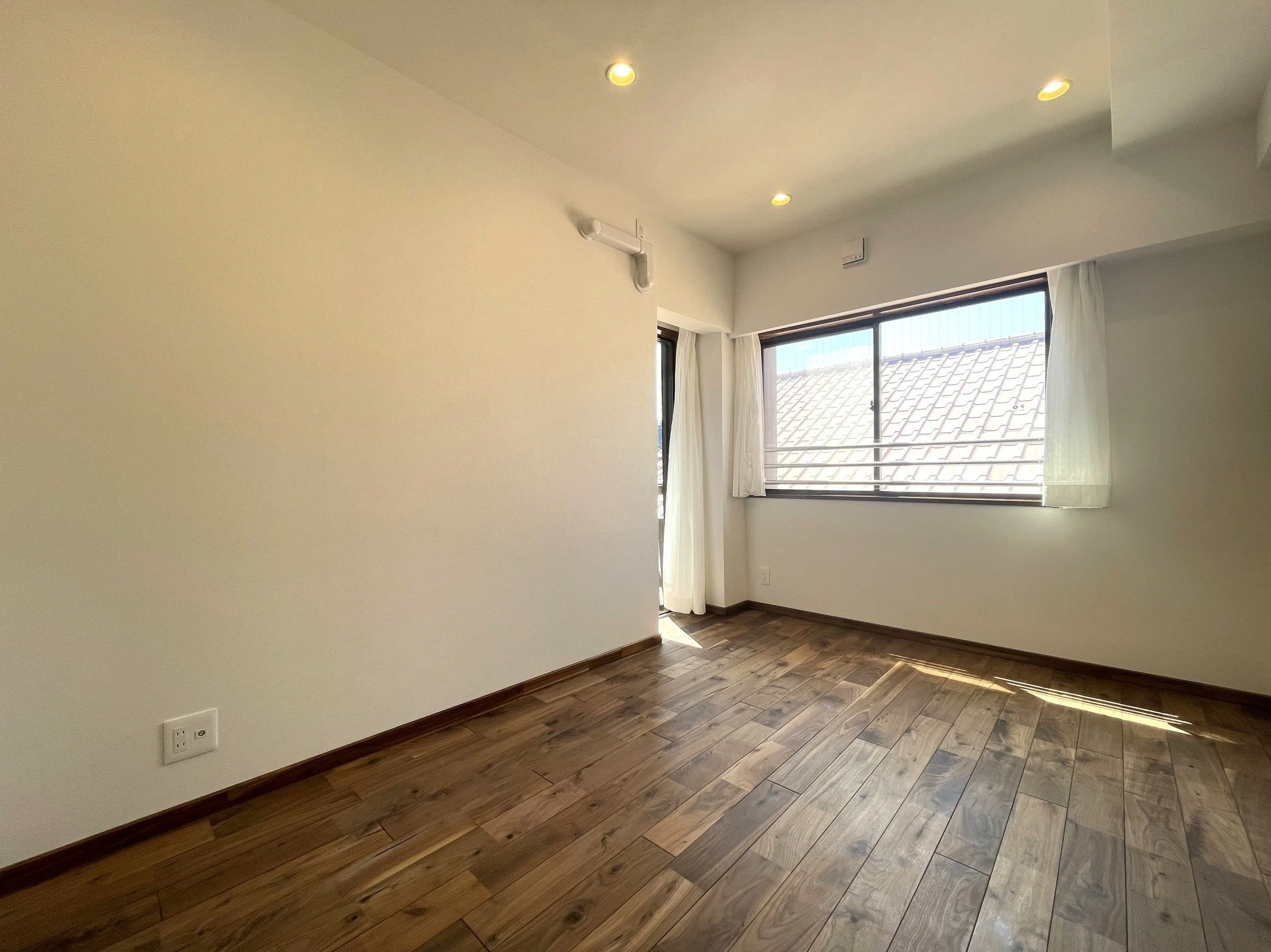 明るい部屋の内装、木製の床に大きな窓と白いカーテン、天井には複数の天井照明。