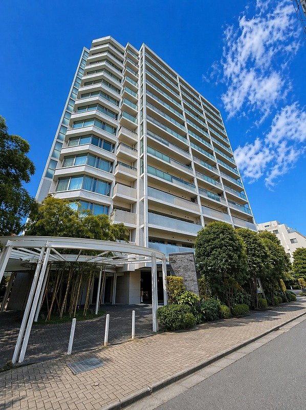 高層の白いマンションと青空。入口に緑の木と歩道がある。