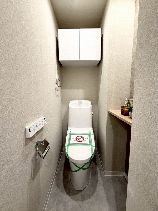 白い便座と壁掛けの収納棚がある狭いトイレで、便座に緑色のテープと禁止のサインが貼られている。