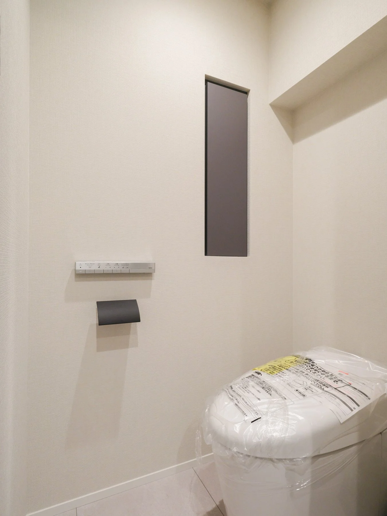 白い壁の部屋のトイレに、新しい便座が紙袋に包まれて置かれている。壁にはリモコンと換気扇のスイッチがある。