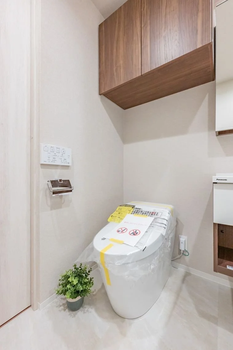 白いトイレと木製の収納棚、トイレットペーパーのホルダー、プラントが置かれたトイレ空間