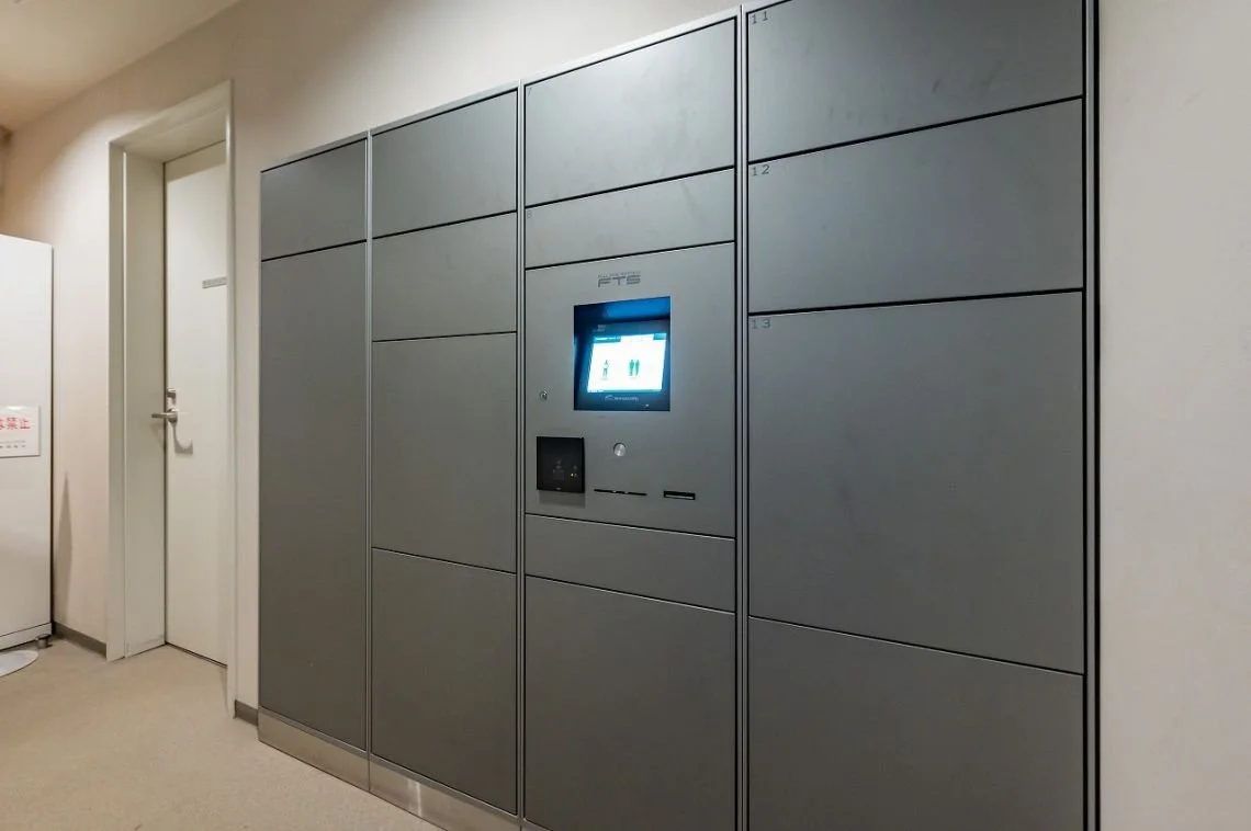 灰色のロッカーとタッチスクリーン式の荷物預かりシステムが設置された室内の壁、左側には白いドアがある。