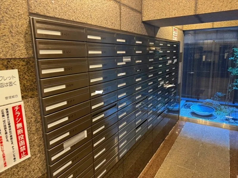 集合郵便箱とその横に貼られた注意書きの掲示板、右側には庭園と水盤のある窓が見える。