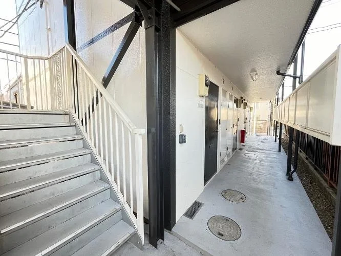 白い建物の廊下と階段の画像。階段は左側にあり、建物の側面に沿って続いている。廊下には複数のドアと電気メーター、排水溝が見える。