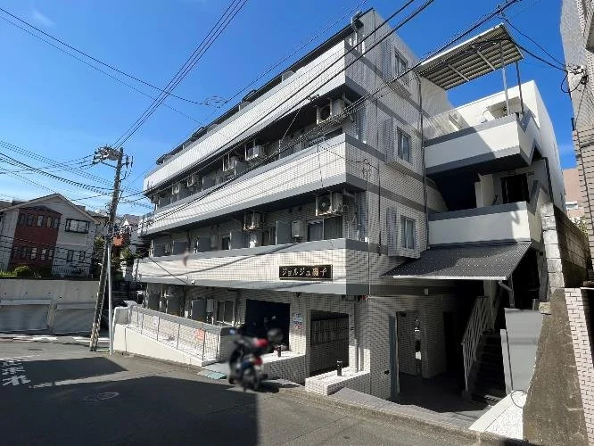 四階建ての白と灰色のアパートメントビル。外にエアコンが設置されており、階段が外側にある。通り側に駐車中のバイクと自動車。