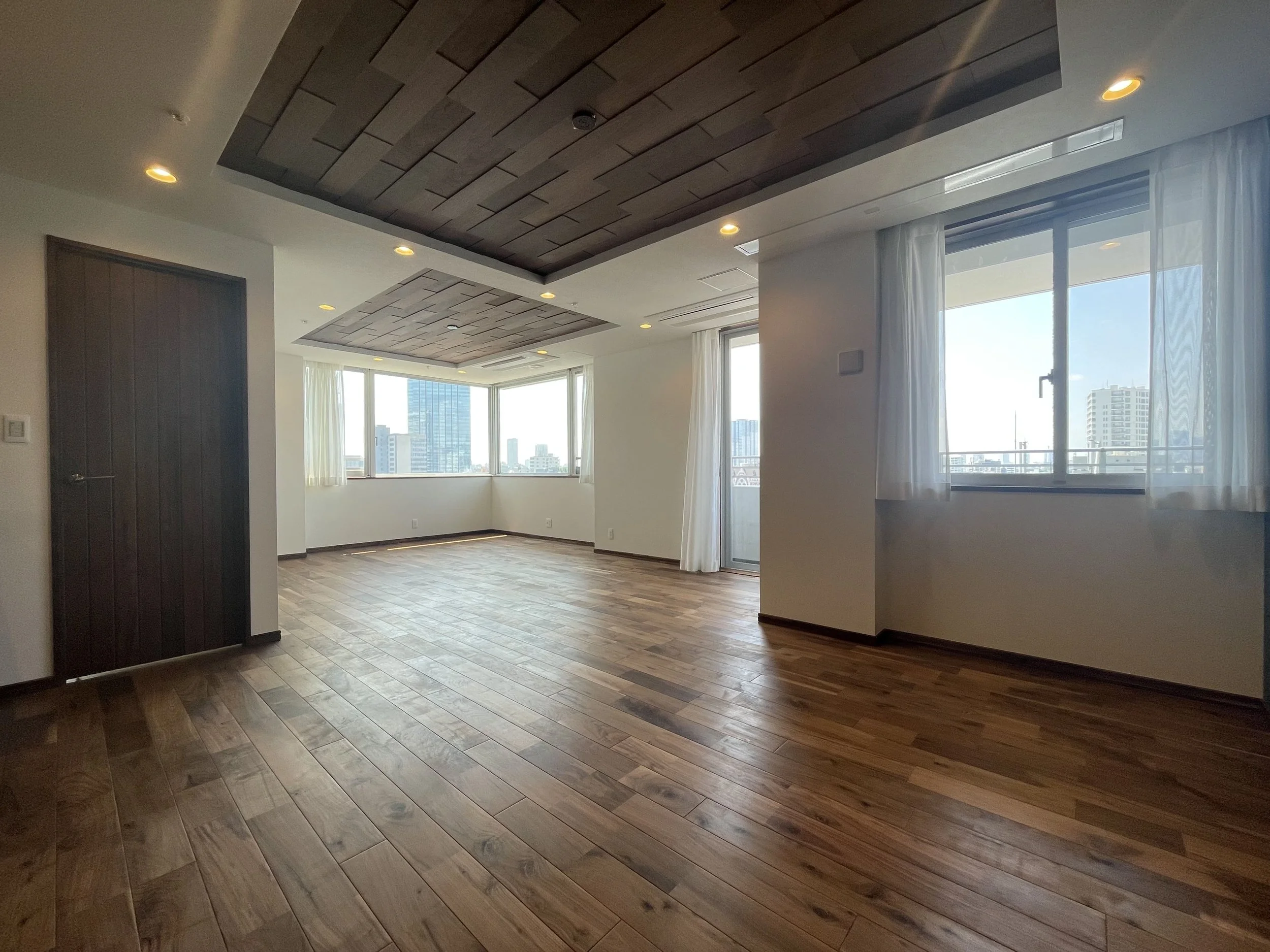 広々とした空間のリビングルームで、大きな窓から自然光が差し込み、都市の高層ビルが見える。木製の床と天井の一部に木目のアクセントがある。