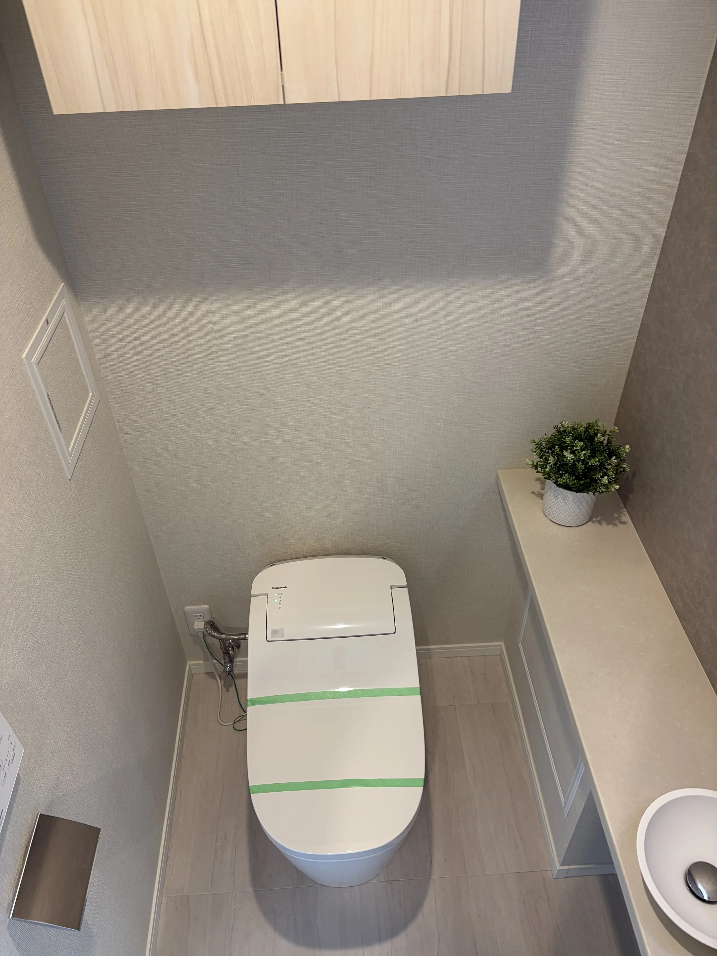 白い便座とトイレットペーパーのホルダー、観葉植物のあるトイレの壁と床の写真