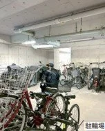 駐輪場に自転車と電動自転車が停められている。後ろにエレベーターやエアコンの室外機が設置された建物の壁が見える。