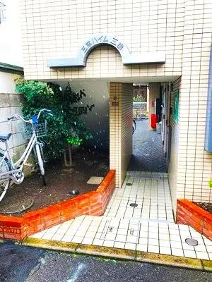 建物の入口付近の様子。自転車と植物があり、タイル張りの通路とレンガの囲いが見える。入口にはアーチ型のサインと記載がある。