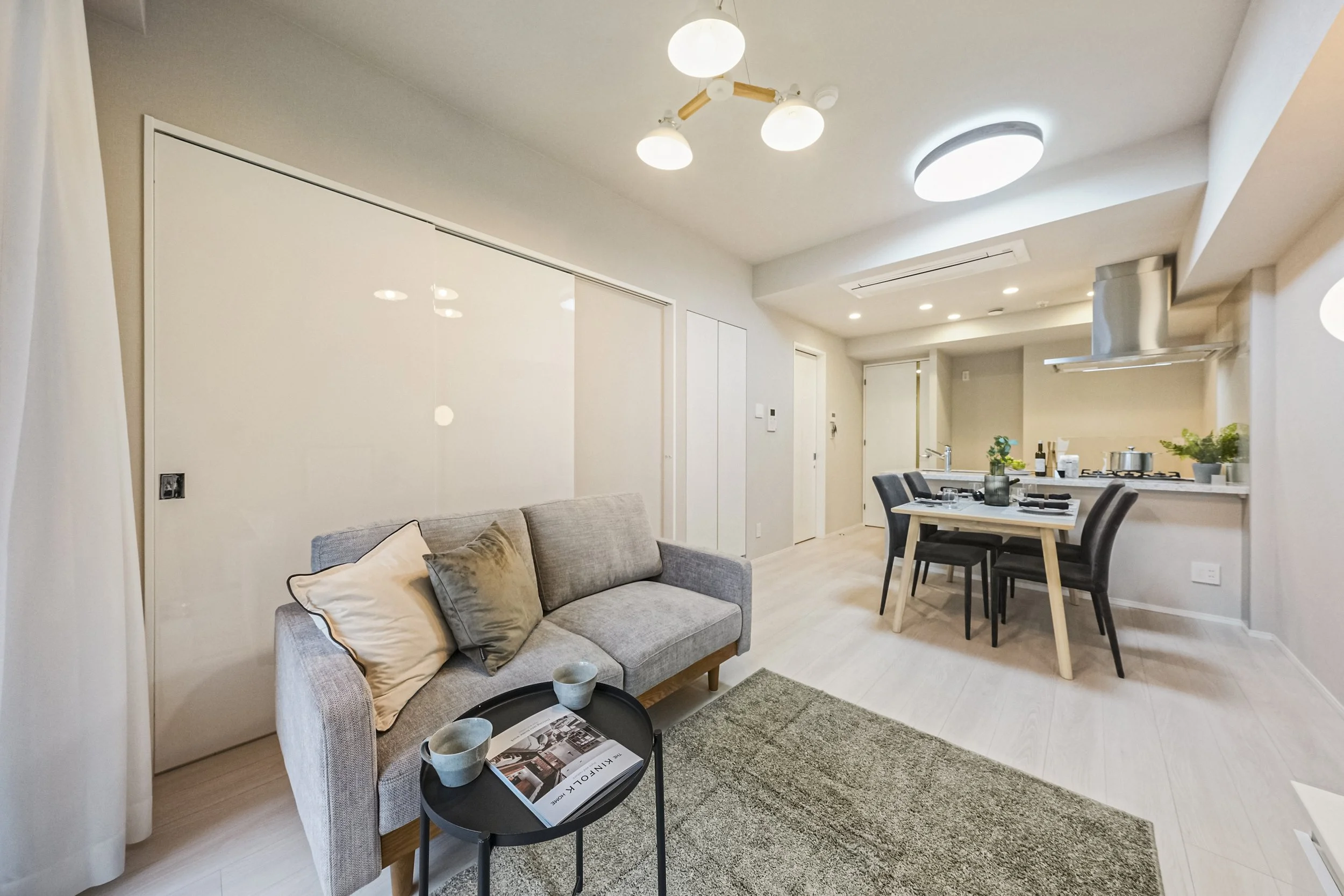 モダンなリビングルームとダイニングキッチンのインテリア風景。灰色のソファと小さな黒いテーブル、ダイニングセット、明るい照明と植物が配置されている。