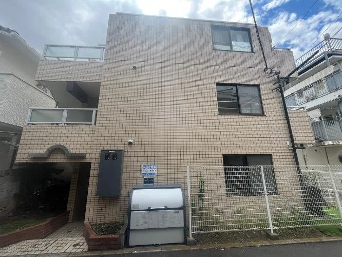 茶色のタイル張りの3階建ての建物。バルコニーと窓がある。前にゴミ箱が置かれ、フェンスで囲われている。