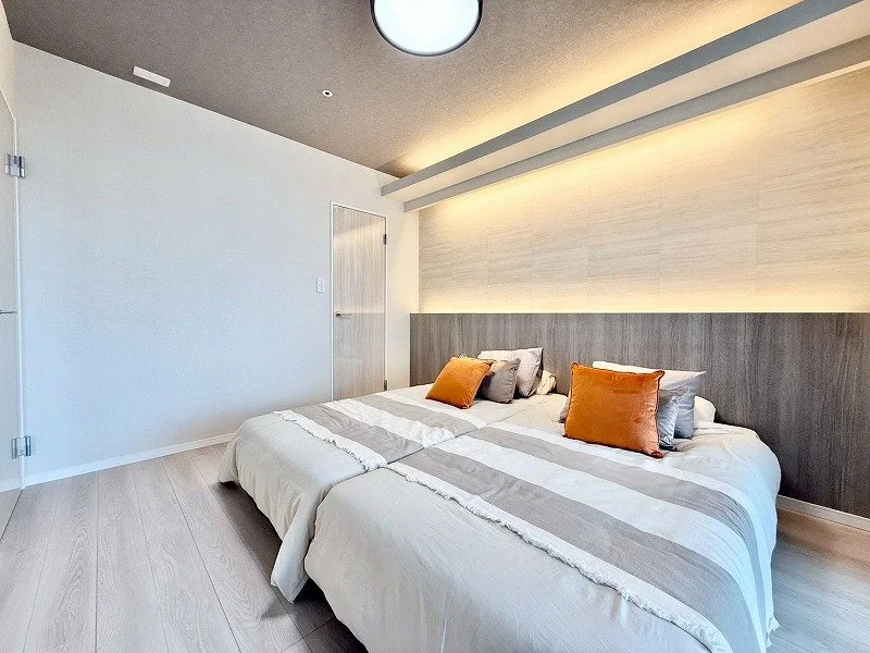 シンプルでモダンなデザインの二つのベッドが並ぶ寝室。壁には間接照明があり、ベッドにはオレンジとグレーの枕が置かれている。