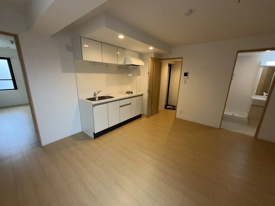 白い壁と木製のフローリングの部屋にキッチンとドアが見える。