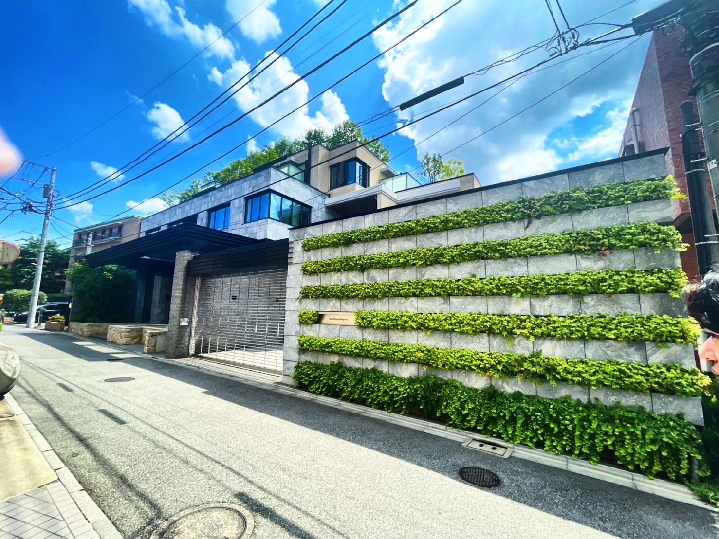 都会の住宅街に建つ現代的なデザインのマンション。外壁には緑の植物が階段状に配置されている。空には白い雲と青空が広がる。