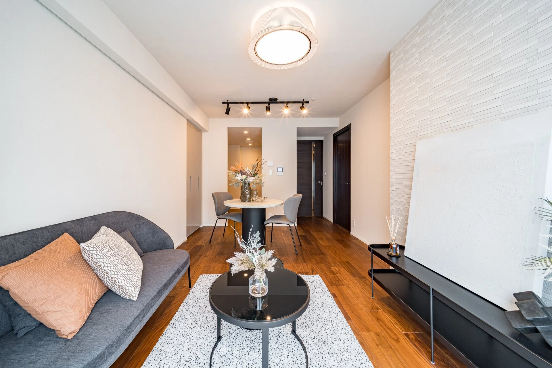 モダンなリビングルームで、グレーのソファとラウンドコーヒーテーブル、白いラグ、白い壁と木製床。丸い天井灯と黒い照明器具、ダイニングテーブルと椅子が見える。