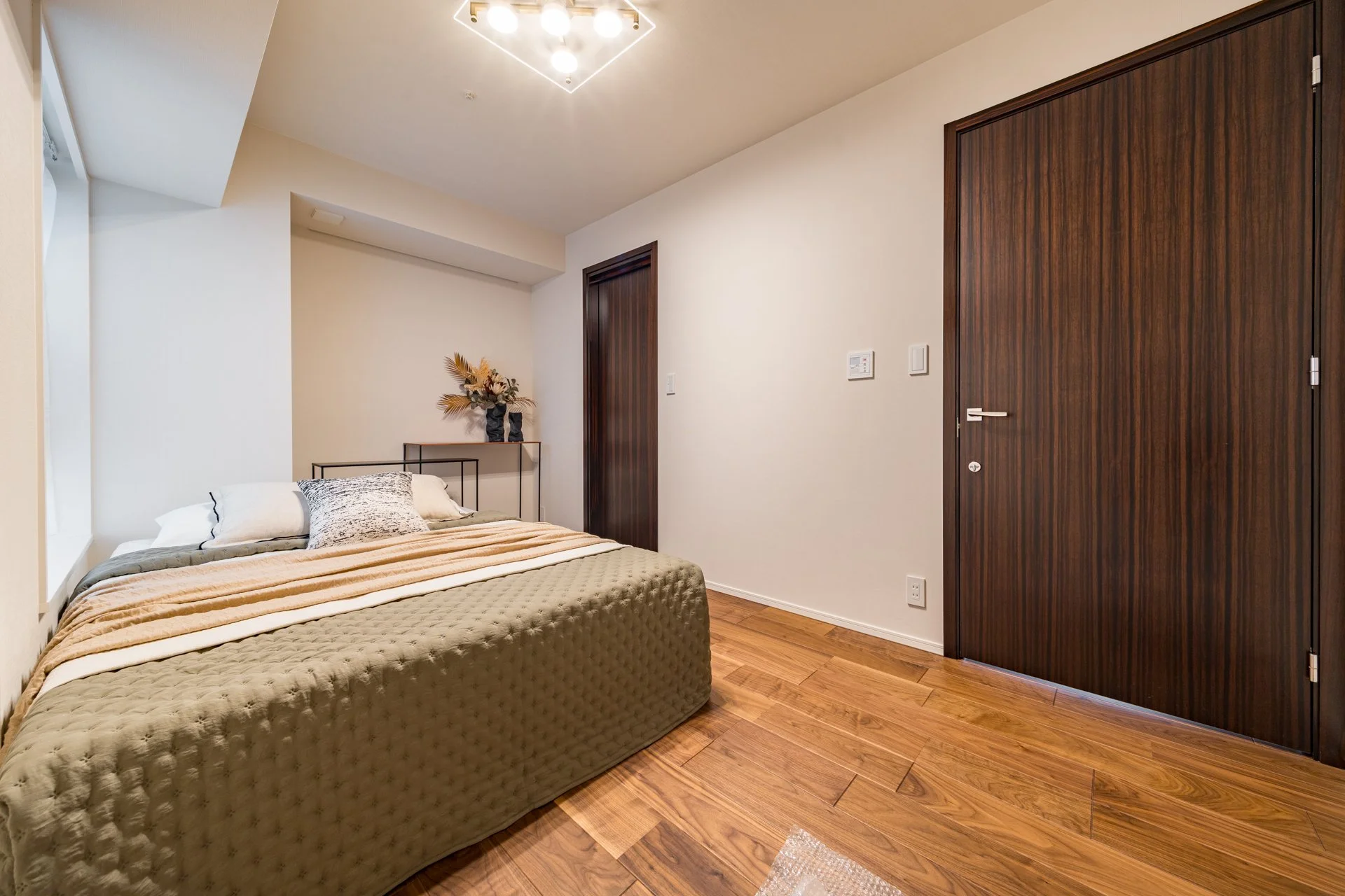 木製の床と木製の扉がある寝室の内装。ベッドには寝具と枕が置かれ、壁の上には照明と飾りと植物が飾られている。