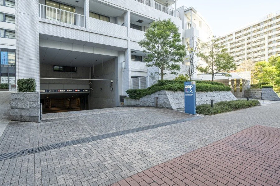 高層の白い建物と舗装された道路、エレベーターの入り口と駐車場の標識、木々と低い生垣が周囲にある都市の景観.