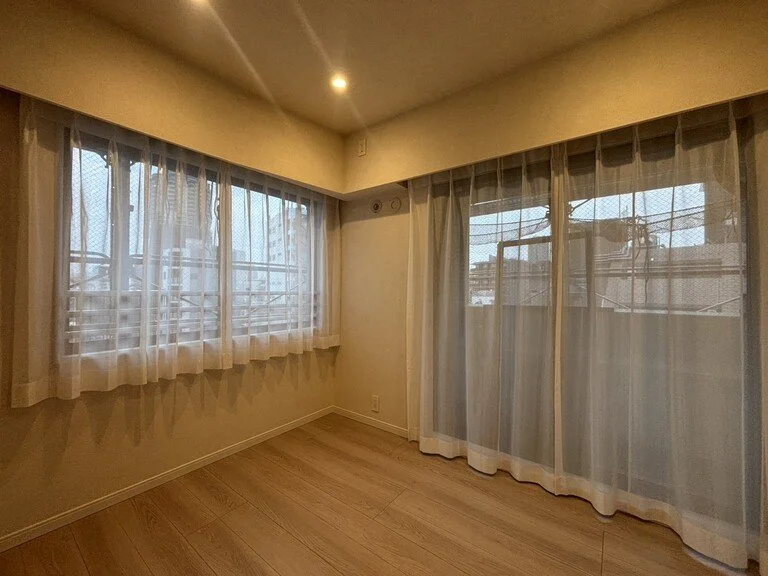 明るい空間の部屋で、壁に大きな窓とカーテンがあり、木製の床が見える