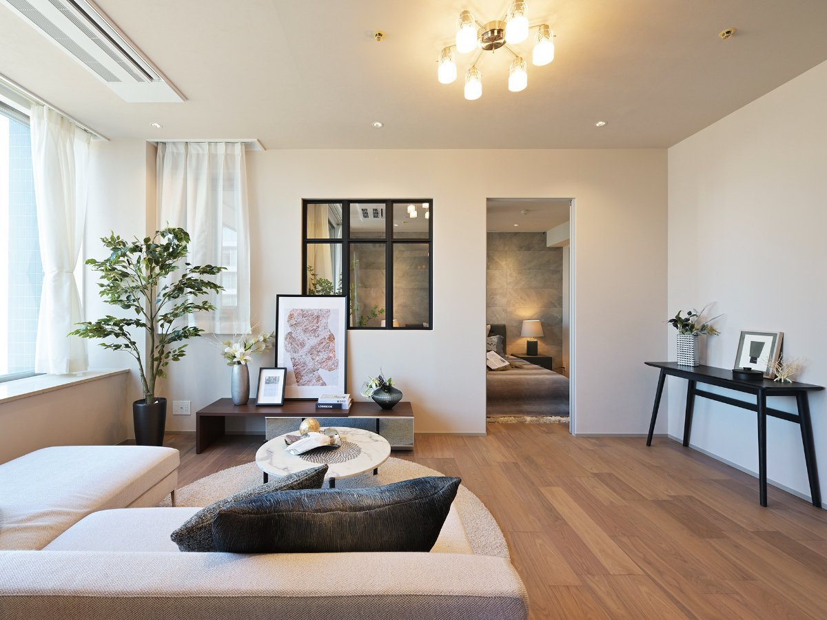 モダンなリビングルーム。ソファ、丸いコーヒーテーブル、観葉植物、壁に飾りとアート、木製のフローリングとシンプルな照明がある。隣に寝室の入り口。