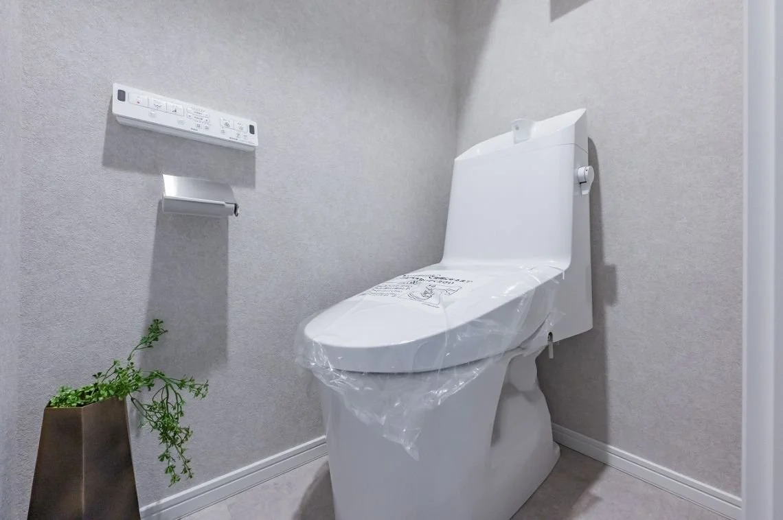新しい洋式トイレとトイレットペーパーのホルダー、壁に取り付けられたリモコン、観葉植物が置かれたトイレ室内の写真