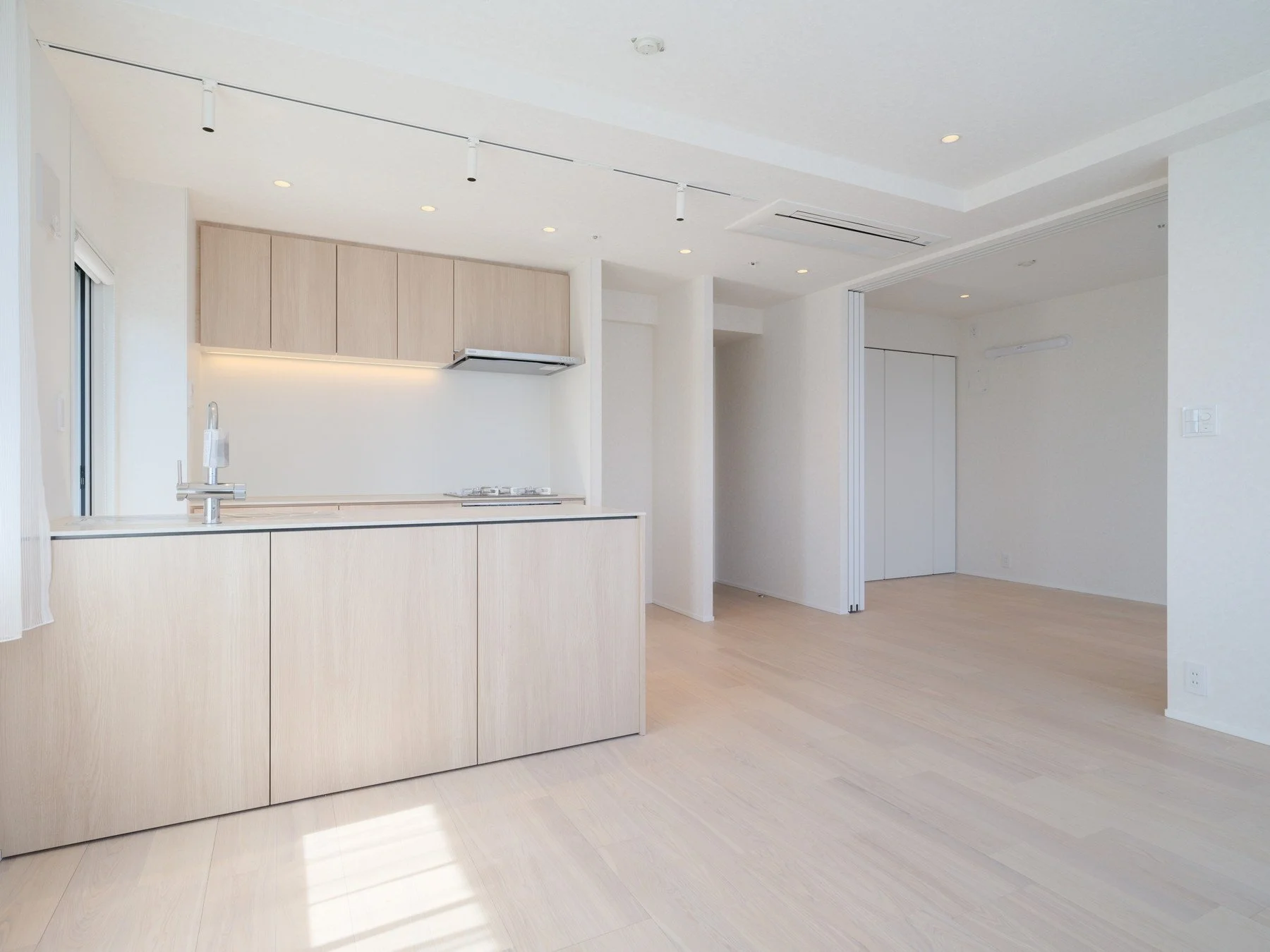 白い壁と天井の無人のキッチンとリビングエリア、木製の床と収納棚。自然光が差し込むシンプルな室内空間。