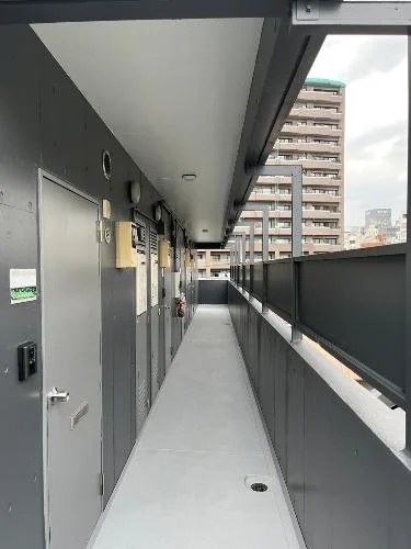 マンションのバルコニーの廊下の画像。左側は扉と設備が並び、右側は手すりと都市の建物が見える風景。
