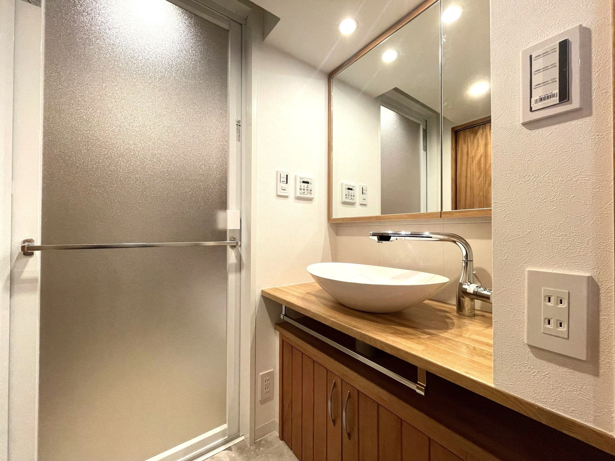 木製のカウンターに置かれたホワイトの陶器製の洗面器と蛇口、背面には大きな鏡とその隣の電気設備のスイッチとコンセントのセットがあり、横の壁には調節可能な温度や湿度を管理する機器が取り付けられている。ドアは金属製で、横に角型のタグがついている。