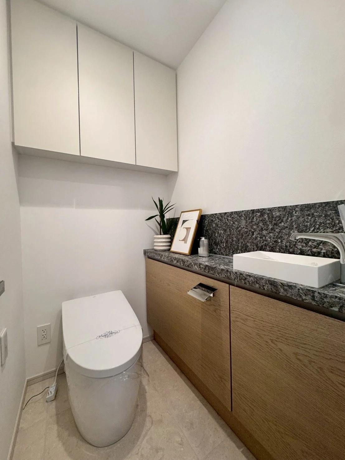 洗面台とトイレがあるモダンなトイレルーム。白い便器、木目調のキャビネット、グレーの大理石風カウンターと背景壁、観葉植物とフレームが飾られている。