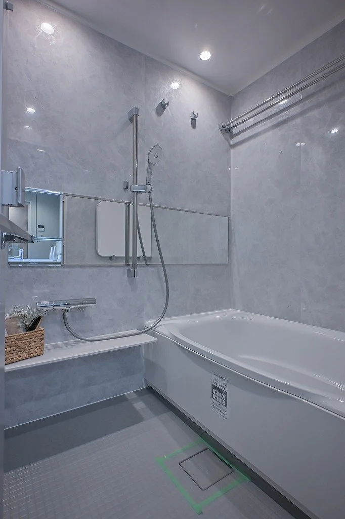 白いバスタブとシャワーヘッドが付いた浴室。鏡とバス用品を置く棚があり、天井には照明と換気扇が設置されている。