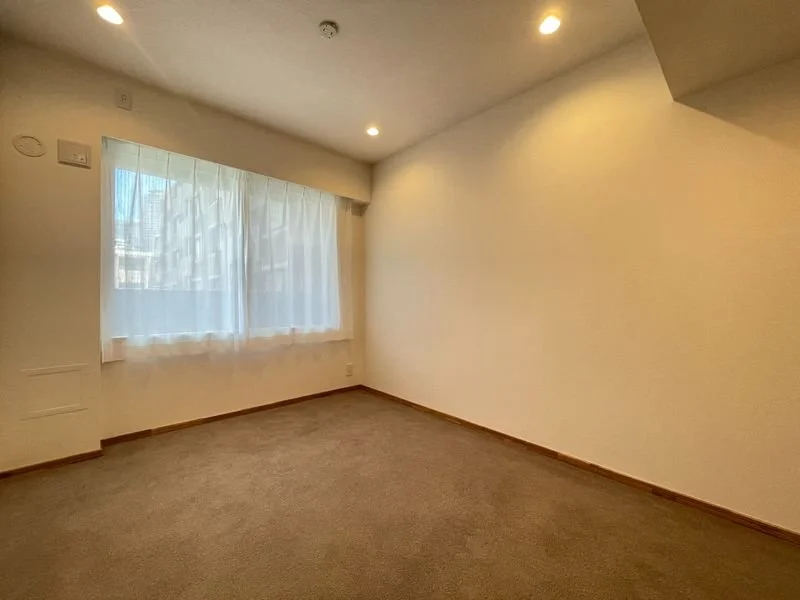 明るい照明のついた空の部屋で、カーテン越しに自然光が入る。窓の外にビルの建物が見える。